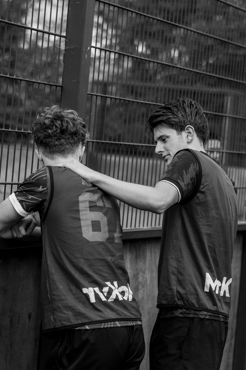 Black and white photo of two players at the pitch fence, one with a hand on the other’s shoulder